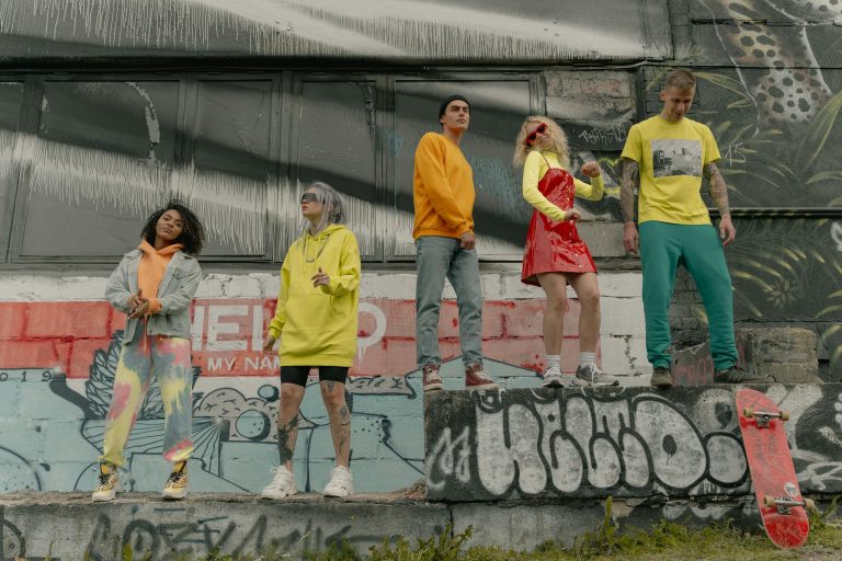 A diverse group of young adults posing stylishly in urban streetwear by a graffiti-covered wall.