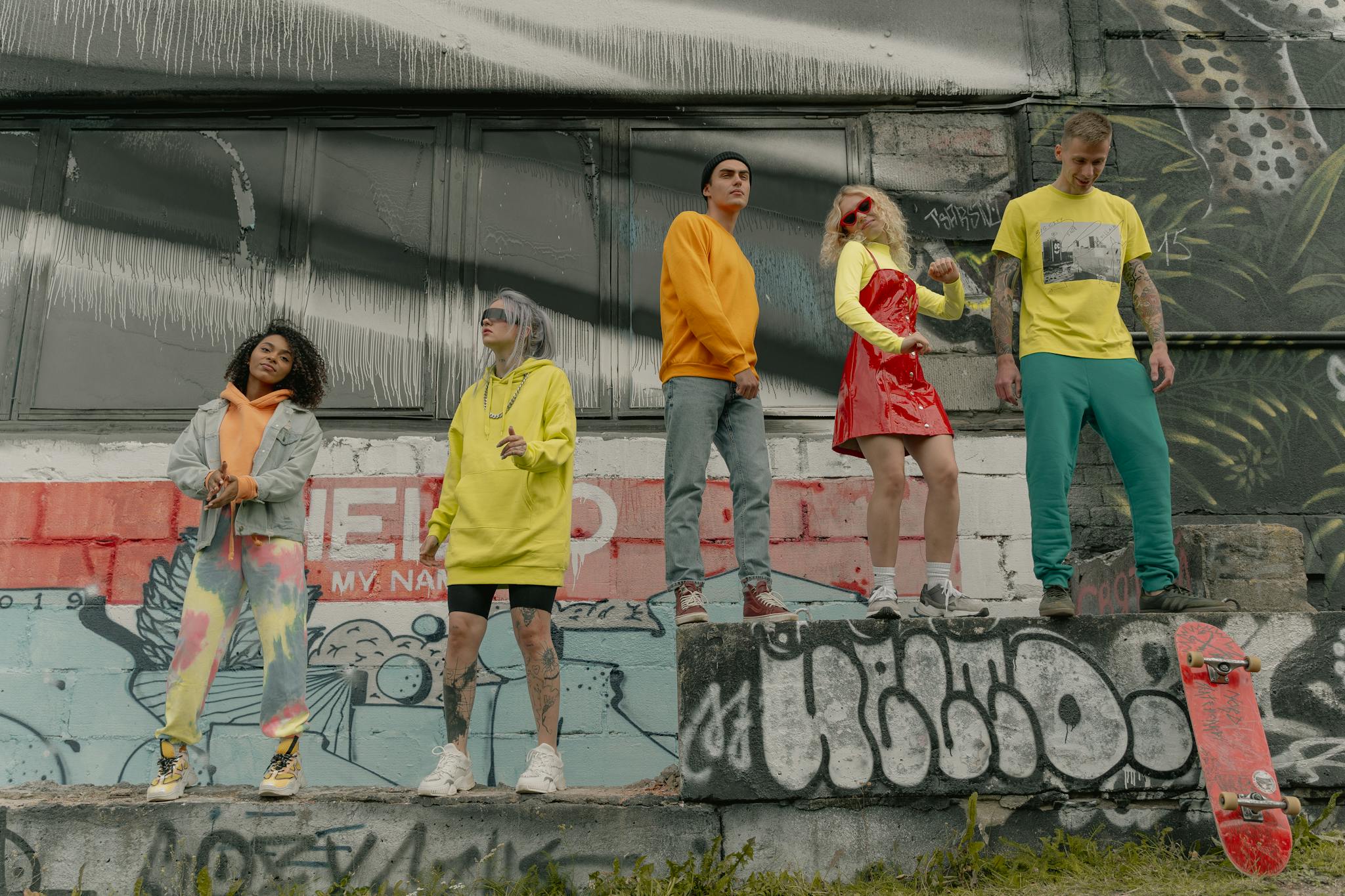 A diverse group of young adults posing stylishly in urban streetwear by a graffiti-covered wall.
