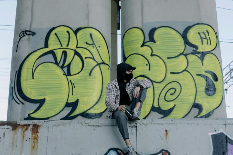 A street artist sits by vibrant graffiti under an urban bridge.