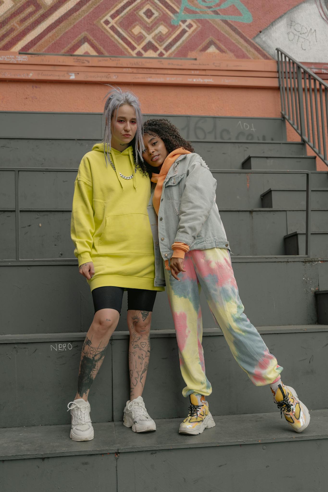 Two stylish women in vibrant urban attire pose on stairs, showcasing tattoos and casual fashion.